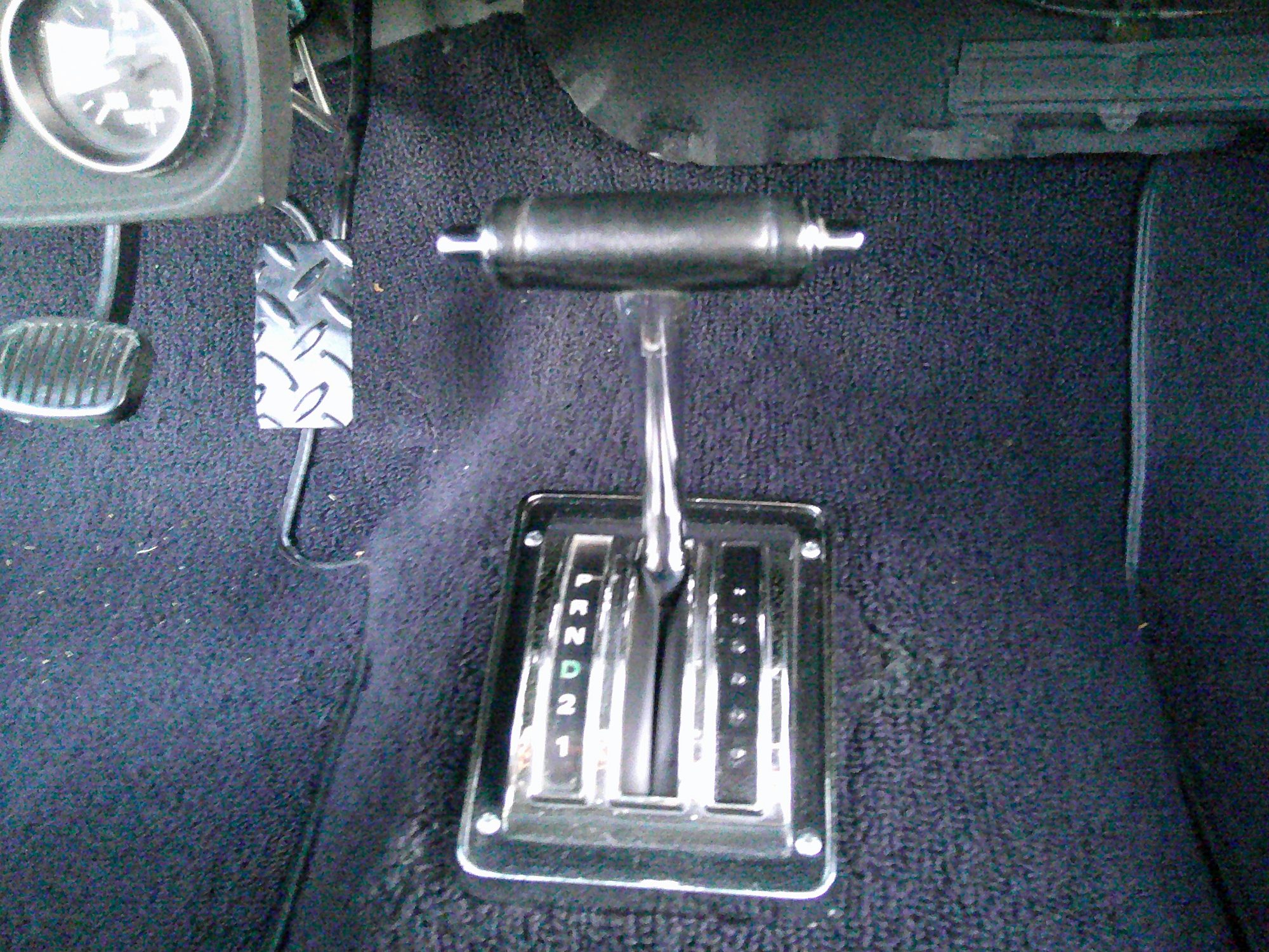 Automatic floor shifter with T-handle and custom pedals over black carpeting.