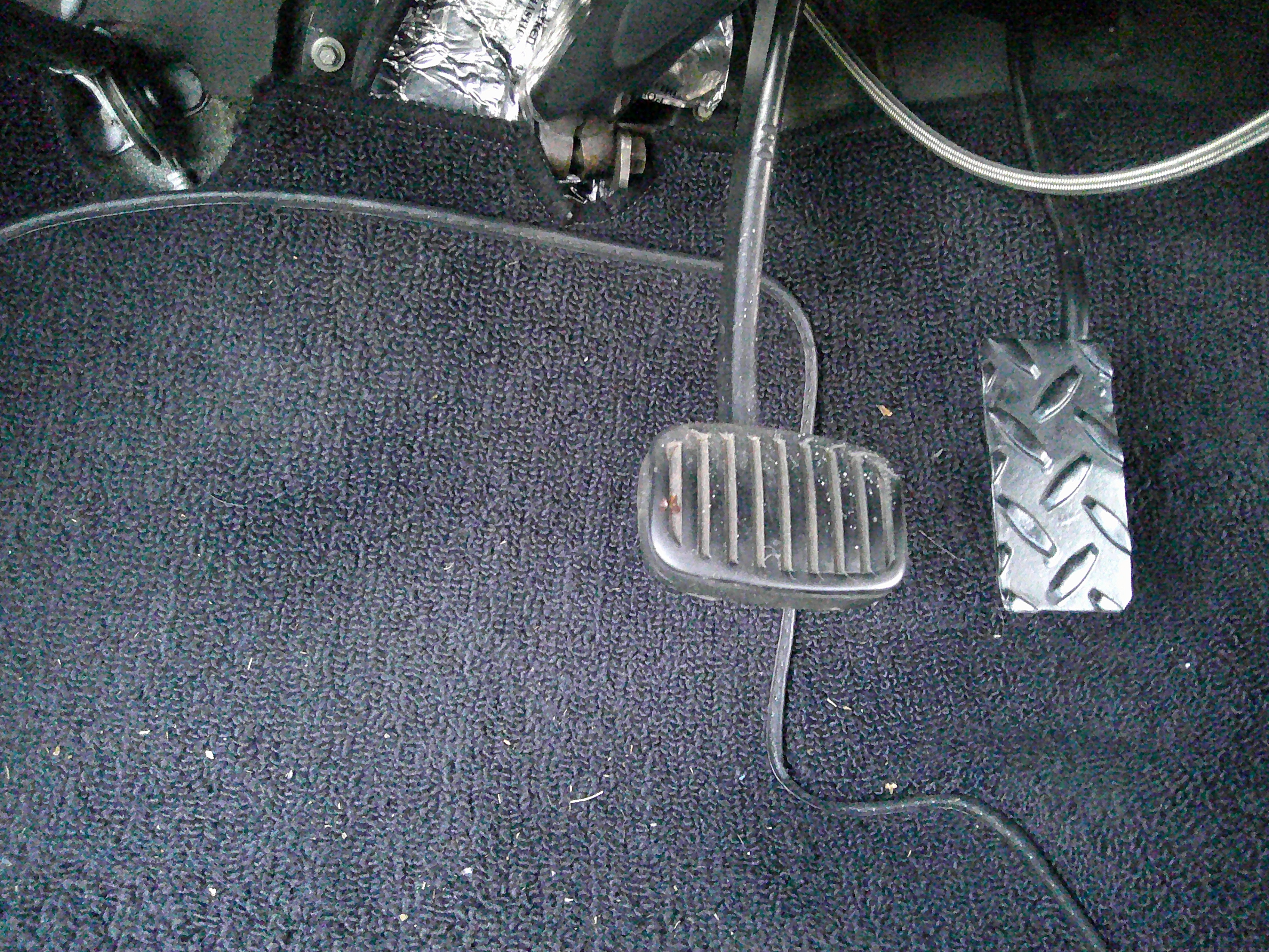 Driver side footwell showing brake and accelerator pedals on black carpeting.