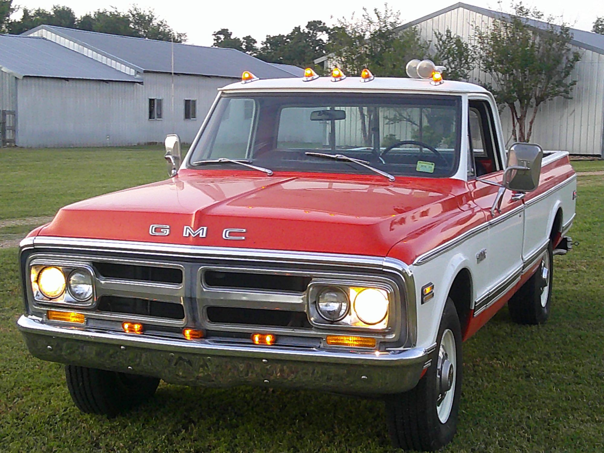Front three-quarter view of a red and white GMC pickup truck with headlights on.